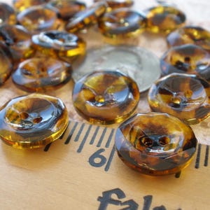 May include: A collection of translucent, tortoiseshell-patterned buttons. The buttons are a mix of amber and brown hues, with a glossy finish. A ruler with inch markings and a coin are also visible.