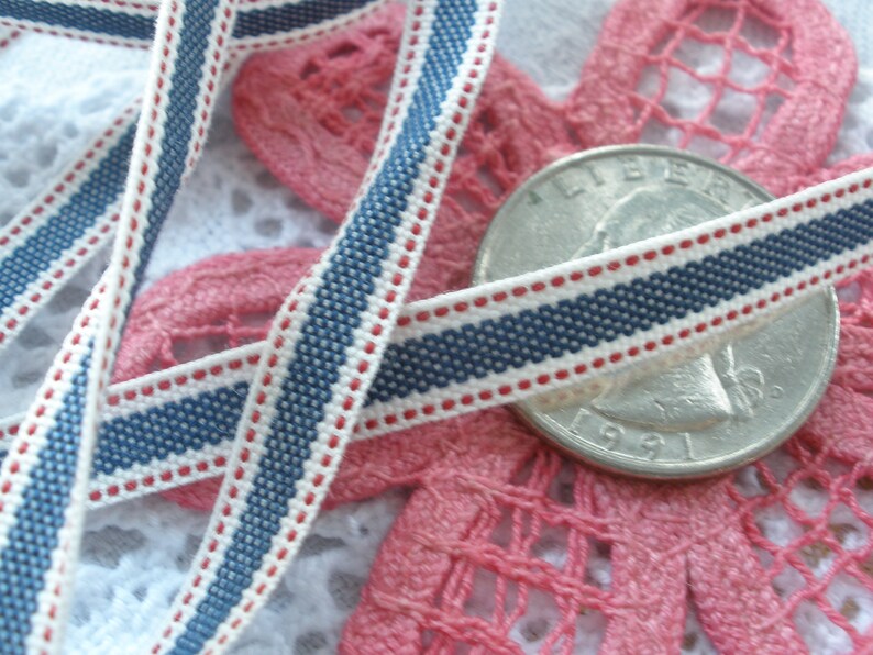 May include: Close-up of a decorative ribbon with a navy blue, white, and red striped pattern. The ribbon is lying on a pink lace doily and a silver coin. The ribbon is approximately 0.5 inches wide.