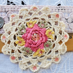 May include: A cream-colored, crocheted doily with a central pink flower design. The flower is surrounded by yellow and green leaves and embellished with sequins. The doily measures approximately 4 inches in diameter.