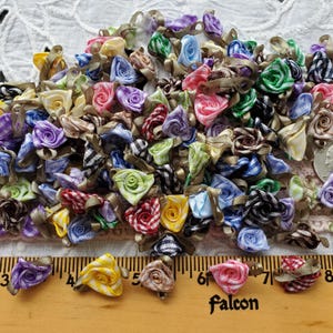 May include: A collection of miniature fabric roses in various colors, including pink, purple, yellow, and blue, with brown ribbon accents. The roses are arranged on a white lace surface, with a wooden ruler marked in inches and the word "falcon" visible.