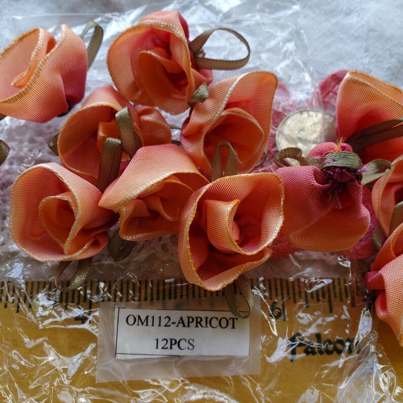 Op de afbeelding: Een close-up van 12 perzikkleurige stoffen rozenbloemversieringen. De bloemen zijn gemaakt van satijnen lint en hebben een licht gerimpelde textuur. De bloemen zijn verpakt in een doorzichtige plastic zak met een wit label waarop "OM112-APRICOT 12PCS" staat.