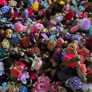 May include: A large collection of colorful fabric flower embellishments. The flowers are made of various fabrics and colors, including pink, purple, blue, green, and yellow. They are arranged in a random pattern and are perfect for adding a touch of whimsy to any craft project.