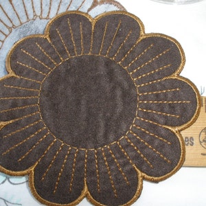 May include: A brown felt coaster with a gold embroidered flower design. The coaster is round and has a scalloped edge.