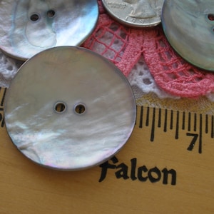 May include: Two iridescent mother of pearl buttons with two holes each. The buttons are round and have a slight sheen. They are sitting on a wooden ruler with the number 5 and 7 visible.