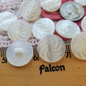 May include: A collection of white, swirl-patterned buttons with a pearlescent sheen. The buttons are arranged on a wooden surface with the word "Falcon" printed on it. A US quarter is also visible.