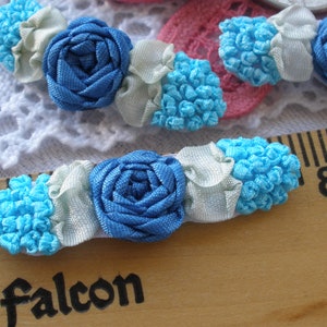 May include: Two blue and turquoise ribbon flower embellishments with white ribbon accents. The embellishments are on a wooden surface with a ruler and the word "falcon" printed on it.