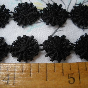 May include: Black lace trim with a repeating pattern of small, round, black rosettes. The trim is measured against a wooden ruler marked with the numbers 4 and 5.