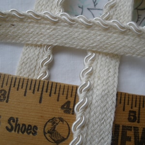 May include: Cream-colored decorative trim with a woven texture and a white, wavy edge. A wooden ruler with the text "Goose Shoes" is in the foreground, measuring the trim. The trim is suitable for sewing and craft projects.