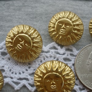 May include: A collection of round, gold-colored metal buttons featuring a raised sun face design with radiating rays. A United States quarter coin is positioned next to the buttons to provide a clear visual reference for their physical size.