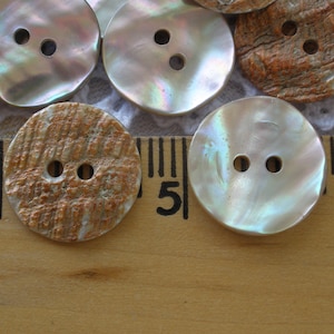 May include: Close-up of several round buttons with two holes each. Some buttons have a pearlescent, iridescent finish, while others have a textured, brown and white appearance. The buttons are arranged on a wooden ruler.