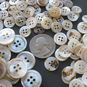 May include: A large pile of white and brown mother of pearl buttons with four holes each. The buttons are various sizes and are arranged in a random pattern. A US quarter is included for scale.