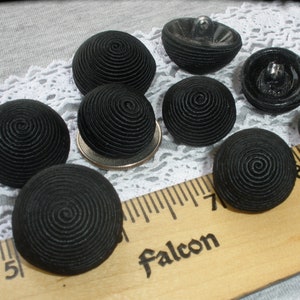 May include: A collection of eight black buttons with a spiral design. The buttons are made of fabric and have a raised, textured surface. The buttons are arranged on a white lace surface with a wooden ruler.
