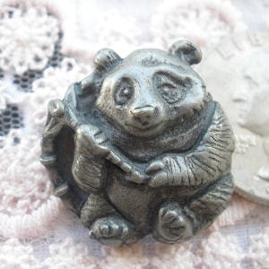 May include: A silver-toned pewter figurine of a panda bear holding a bamboo stalk. The panda is sitting upright with its legs crossed and has a friendly expression.