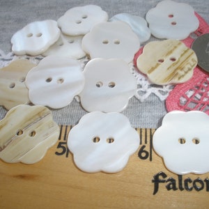 May include: A collection of white and beige flower-shaped buttons with two holes each. The buttons are arranged on a gray fabric background with a ruler and a quarter for scale.