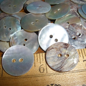 May include: A collection of iridescent mother-of-pearl buttons with two holes. The buttons are various shades of gray, white, and brown. Some buttons have a marbled pattern. The buttons are arranged on a wooden surface with a ruler.