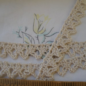 May include: Close-up of two strips of cream-colored crochet lace with a scalloped edge. The lace is lying on a white background with a floral pattern in yellow, blue, and green. A wooden ruler is at the bottom.