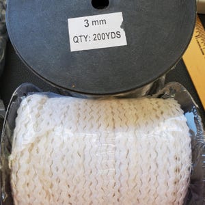May include: A spool of black ribbon with a label that reads "3 mm QTY: 200YDS." Below the spool is a package of white, wavy trim. The trim is wrapped in clear plastic.