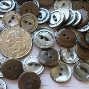 May include: A collection of vintage buttons made from mother of pearl. The buttons are in various shades of brown and blue with a iridescent sheen. Some buttons have two holes, while others have four holes. The buttons are arranged on a white surface with a quarter for scale.