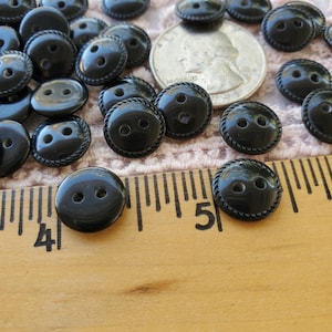 May include: A collection of glossy black buttons with two holes and a rope-like edge. A ruler shows the buttons are approximately 0.75 inches in diameter. A US quarter is also present, providing a size reference. The buttons are on a textured surface.