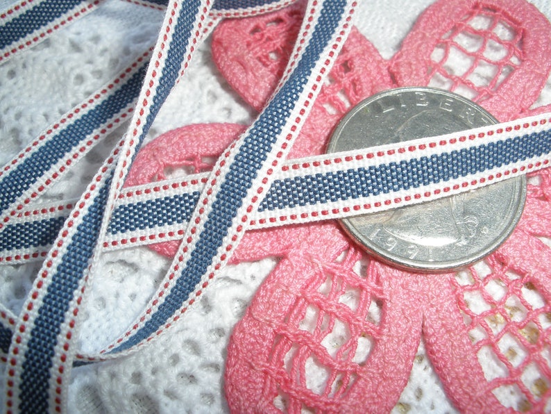May include: Close-up of a blue, white, and red striped ribbon. The ribbon is approximately 0.5 inches wide and is lying on a pink floral lace design. A US quarter is also in the image.
