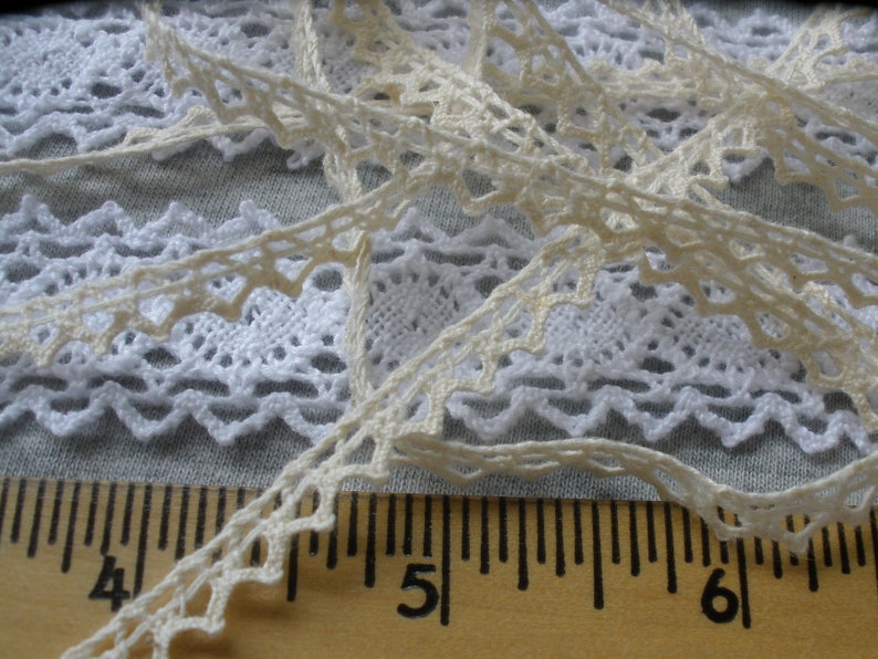 May include: Two pieces of white lace trim with a scalloped edge. The lace is laid out on a wooden ruler with markings for inches. The ruler shows the numbers 4, 5, and 6.