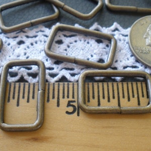 May include: A close-up of several antique brass D-rings, measuring approximately 1 inch in length, against a white lace background and a wooden ruler.