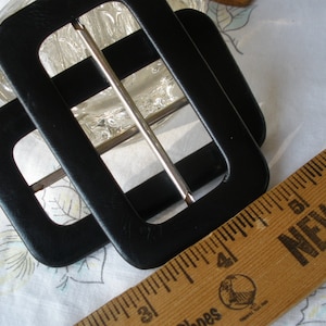 May include: Two black rectangular belt buckles with silver-colored bars. A wooden ruler with the text "NEW TO" is in the frame. The buckles are on a floral patterned fabric.