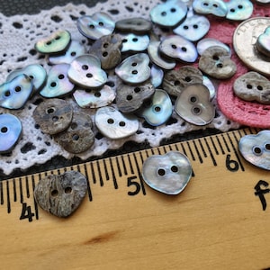 May include: A collection of iridescent heart-shaped buttons with two holes each. The buttons are made of mother of pearl and have a natural, rustic look. The buttons are scattered on a white lace fabric and a wooden ruler with inch markings.