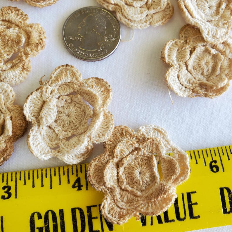 May include: Close-up of several crocheted flower appliques in shades of beige and tan. A US quarter coin is next to the flowers for size comparison. A yellow ruler is in the foreground, with the word "GOLDEN" visible.