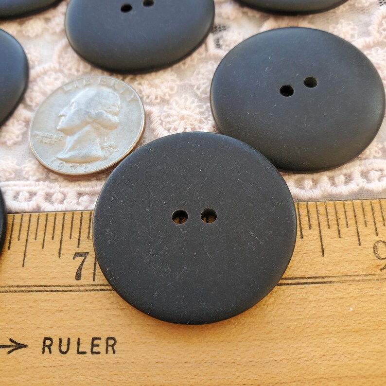 May include: A collection of large, matte black buttons with two holes each. A silver quarter is next to the buttons for size comparison. The buttons are on a wooden surface with a ruler.