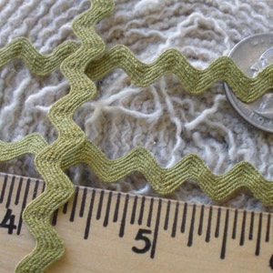 May include: Olive green zigzag ribbon, a decorative trim, is shown against a textured, off-white background. A ruler and a coin are also in the image, providing scale. The ribbon's wavy design adds a unique touch to craft projects.