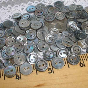 May include: A large pile of gray and iridescent mother of pearl buttons. The buttons are all round and have two holes. They are arranged in a random pattern on a white surface. A ruler is visible in the bottom right corner of the image.