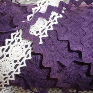 May include: Close-up of purple and white decorative trim. The purple trim has a wavy, textured design. White, crocheted lace with a circular pattern is also visible. This trim is suitable for various craft projects.