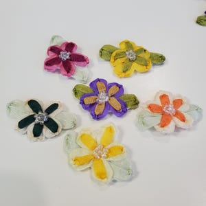 May include: A collection of colorful, fabric flower embellishments. Each flower features a central beaded accent and is composed of petals in shades of pink, yellow, purple, orange, and green. The flowers are arranged on a white surface.