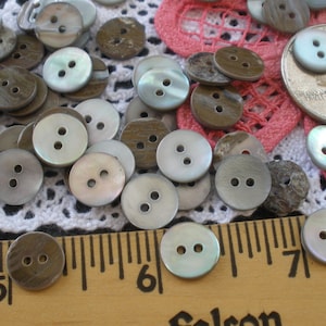 May include: A collection of 2-hole shell buttons in various shades of brown and gray. The buttons are arranged on a white crocheted surface with a pink crocheted surface in the background. A ruler is in the foreground with the numbers 5, 6, and 7 visible.