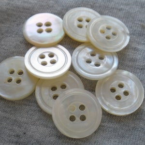 May include: A collection of ten white pearl buttons with four holes each. The buttons are arranged in a circular pattern on a light gray fabric background.