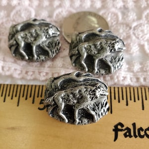 May include: Three silver-toned metal buttons, each featuring a detailed relief of a wolf standing in tall grass. The buttons have a slightly weathered appearance, suggesting an antique or vintage style. A ruler and a coin are also in the image.