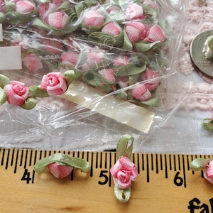 May include: Pink and white satin ribbon roses with green leaves. The roses are in a clear plastic bag and also arranged on a wooden ruler. The roses are approximately 0.5 inches in diameter.