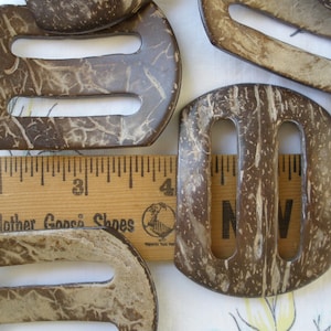 May include: A set of five brown coconut shell belt buckles with two holes each. The buckles have a natural, rustic look with a light brown color and a white marbled pattern.