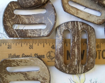May include: A set of five brown coconut shell belt buckles with two holes each. The buckles have a natural, rustic look with a light brown color and a white marbled pattern.