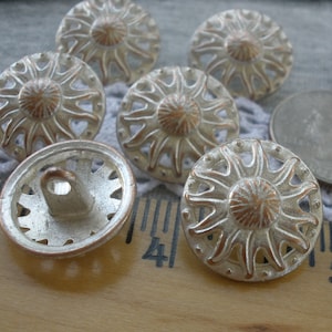 May include: A collection of ten silver-toned buttons with a sunburst design and a copper-colored center. The buttons have a raised rim and a hole in the center for sewing.