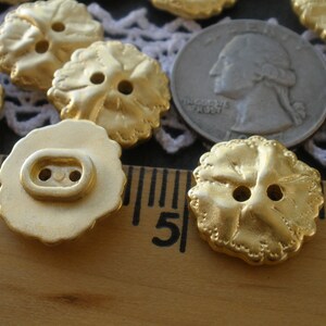 May include: A close-up of ten gold-colored metal buttons with a flower design. The buttons are arranged in a semi-circle on a wooden surface with a ruler.