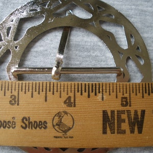May include: A silver-toned metal belt buckle with a decorative, openwork design. The buckle measures approximately 4 inches in diameter.