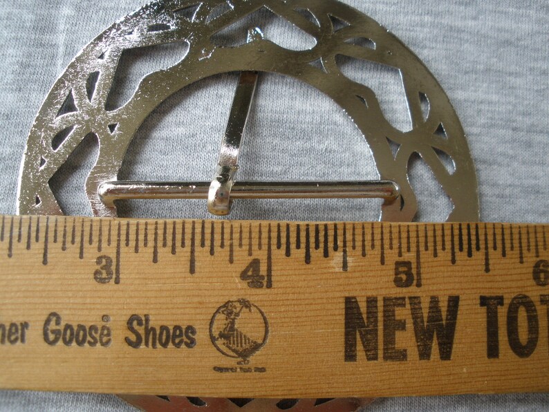 May include: A silver-toned metal belt buckle with a decorative, openwork design. The buckle measures approximately 4 inches in diameter.