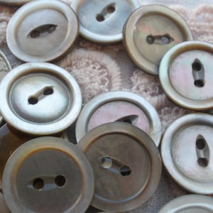 May include: A collection of round, iridescent buttons in shades of gray, brown, and white. Each button has two holes for sewing. The buttons vary in size and are arranged on a textured, light pink surface.