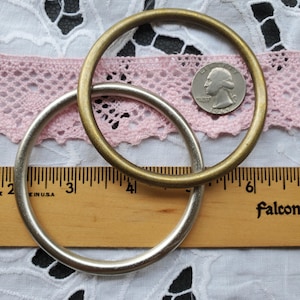 May include: Two metal rings, one silver and one brass, are shown on a wooden ruler with a pink lace background. The silver ring is 3 inches in diameter and the brass ring is 4 inches in diameter.
