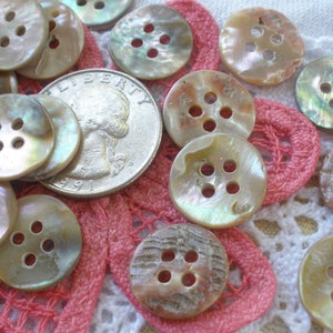 May include: A collection of iridescent mother-of-pearl buttons with four holes each. The buttons are various shades of cream, beige, and brown. They are arranged on a white lace doily with a pink crocheted border.