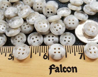 May include: A close-up of a group of white buttons with a gray marbled pattern. The buttons have four holes each and are arranged in a random pattern. A ruler is visible in the bottom of the image, with the numbers 5, 6, and 7 visible. The word "Falcon" is printed on the ruler.
