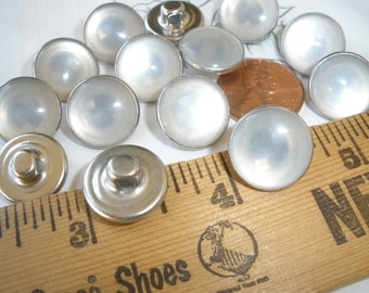 May include: A collection of silver-colored, domed buttons with a pearlescent sheen. The buttons are arranged on a white surface with a wooden ruler and a penny for scale. The buttons have a metal shank on the back.