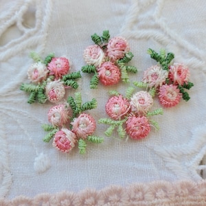 May include: Five embroidered flower clusters with pink and white petals and green leaves. The flowers are stitched on a white fabric background.